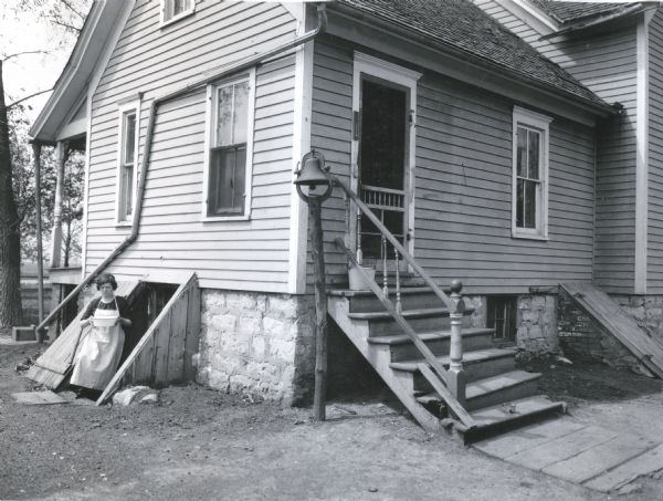 A woman wearing an apron and holding a bowl is posing on the stairs of a cellar bulkhead on the side of a farmhouse. A bell on a wooden pole is next to the back steps leading into the house. The corner of a porch is on the left side. Another cellar bulkhead is on the right.
