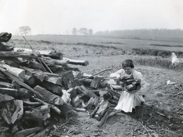 A woman, possibly Anna Neary, gathering pieces of chopped wood in her arms as she kneels near a wood pile on the A.E. Bancroft farm.