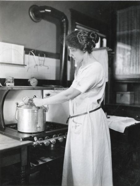 Mrs. Waggoner opening a pressure cooker on a stove in a farmhouse, possibly as part of a home canning process.