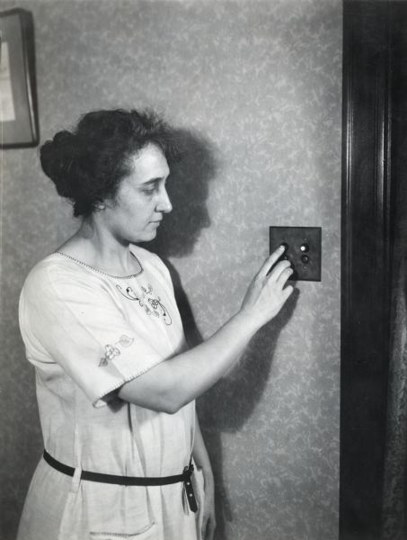 Mrs. Waggoner pushing a button on an electric light switch on the wall in her farmhouse.