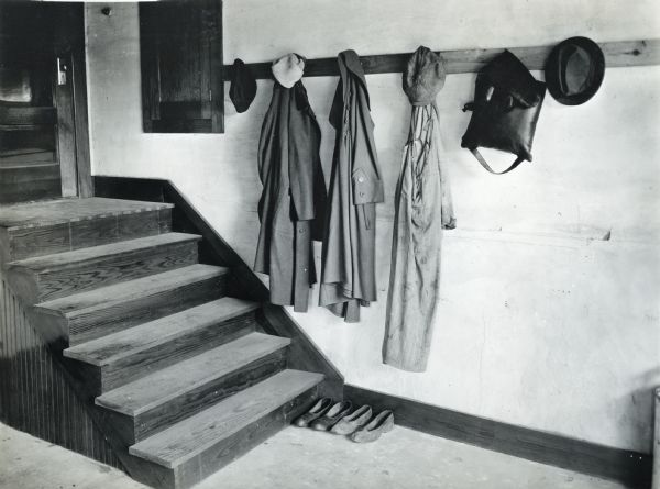 Coats, hats, and bags hanging from a coat rack inside a farmhouse entryway at the home of Adolph Betz. Two pairs of shoes are lined against the wall at the base of a set of wooden stairs.