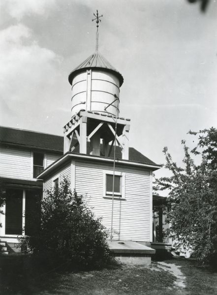 A water supply tank standing atop what appears to be a shed in the back of a farmhouse. A weather vane is at the top of the water tank.