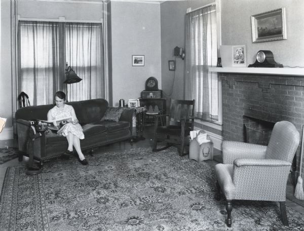 Mrs. L.A. Hawkins sitting on a couch while reading "The Farmer's Wife" magazine in the living room of her home. A fireplace is on the opposite wall, and chairs, a radio, and other furniture are arranged around the room.