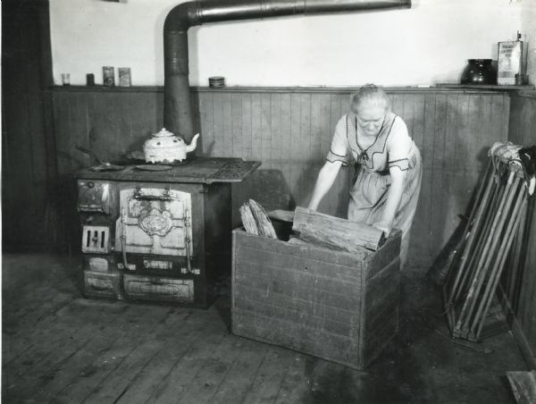 A woman moving a box of firewood inside a farmhouse. A teakettle is standing on a stove to the left.