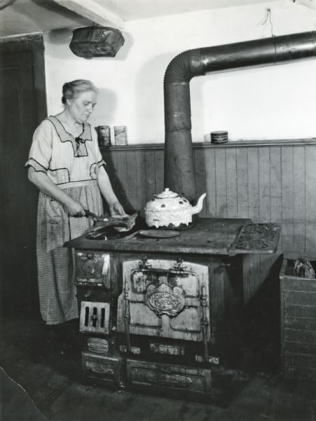 Mrs. John Esche using a woodburning stove marked "Home Comfort" while preparing food. A crate holding firewood is to the right of the stove.