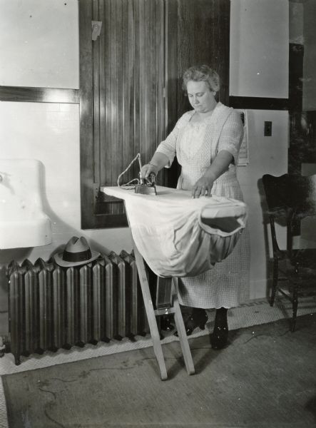 Mrs. Betz ironing a skirt on a wall-mounted foldout ironing board. A radiator is along the wall near a sink, and a wooden chair is on the right.