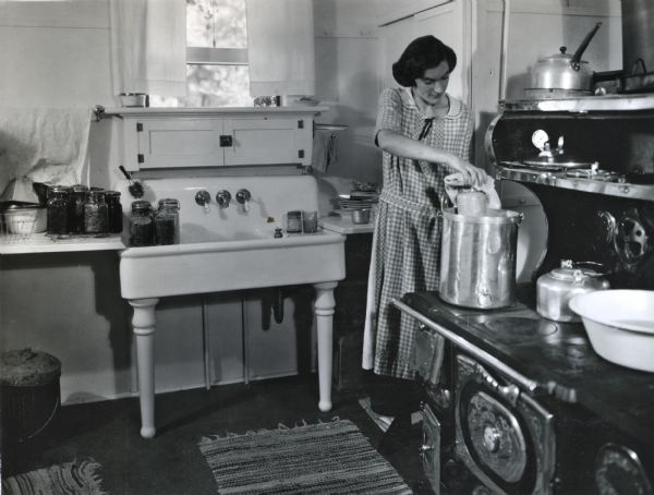 Margaret Monier using a pressure cooker while canning in the home of Miss Marilla Zearing. Cans of produce are cooling on wire racks on the counter and in the sink in the background.