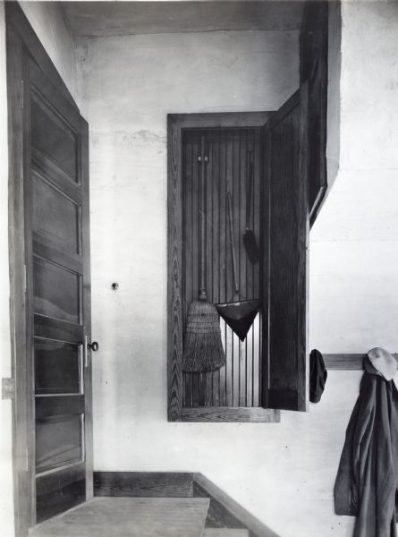 A broom, dustpan, and a duster hanging in a cleaning supply closet at the top of a staircase in the house of Adolph Betz.
