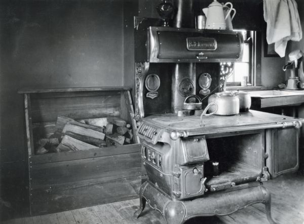 A wooden storage box for firewood behind a woodburning stove in a farmhouse kitchen. A sink with a hand-powered water pump is on the right.