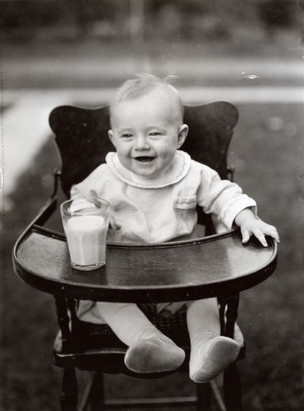A baby sitting in a wooden highchair with a glass of milk placed on the tray.