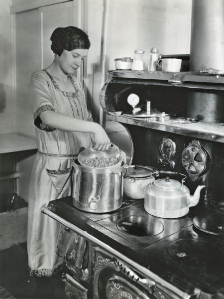 Miss Larson using a pressure kettle to cook on a wood-burning stove in a farmhouse kitchen.