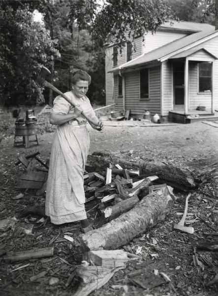 A woman wearing a dress using an axe to split a log into firewood. A hand-operated washing machine is standing in the yard of a farmhouse in the background.