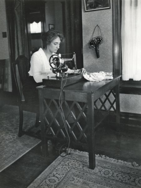 A woman using an electric sewing machine while sitting at a table with latticed sides.