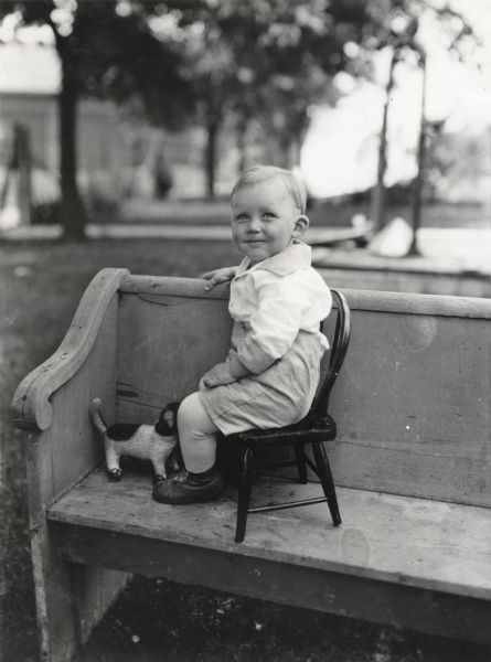 A boy is sitting on a wooden chair placed on top of an outdoor wooden bench. A toy dog with wheels on its legs is on the bench beside him.