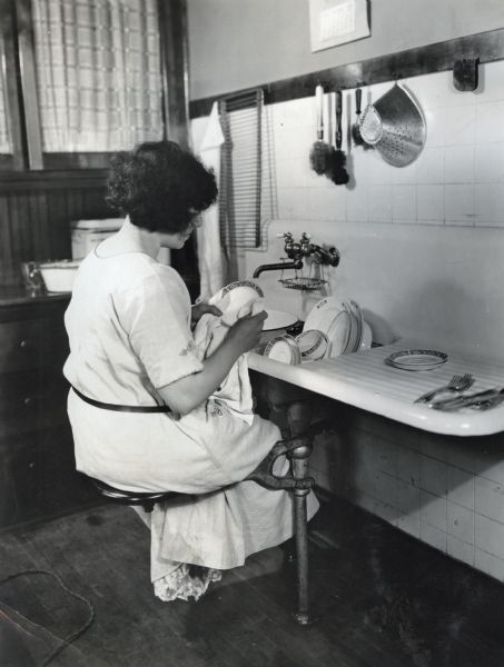 Mrs. Guener washing dishes while sitting on a stool attached to a sink in J.O. Waggoner's home.
