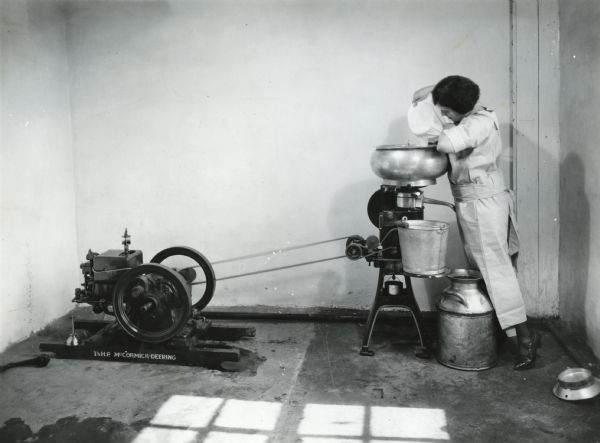 A woman using a cheesecloth to strain the contents of a bucket into a cream separator at International Harvester's Hinsdale experimental farm (Harvester Farm). The separator is powered by a 1.5 horsepower McCormick-Deering engine.