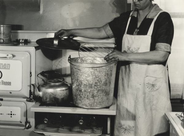 Woman in kitchen demonstrating the canning process. Original caption reads, "pail and wire rack for cold pack canning." The photo was taken at Harvester Farm.