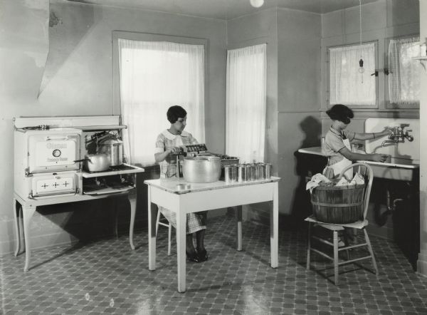 Woman and young girl working in a kitchen canning beans. The girl is standing at the sink running a faucet while the woman is seated at a table using a bean cutter. Original caption reads, "good stove, good light, working space and good sink."
