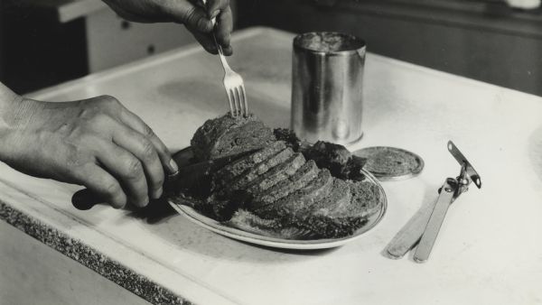 Close-up of a person's slicing a plate of canned meatloaf. Original can and can opener are on the table next to the food.
