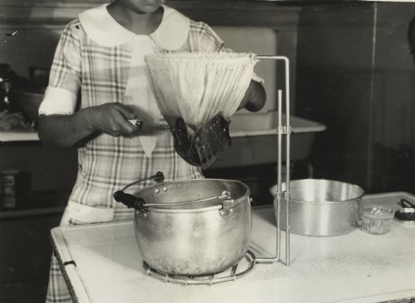 Woman working in kitchen straining jelly bags over pot in preparation for canning. The photograph was taken at an "IHC Farm," probably in Hinsdale, Illinois.