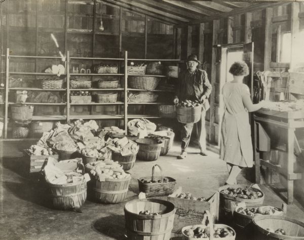 Man carrying baskets of field crops in to the receiving room of a community kitchen. A woman wearing an apron is standing near the door.