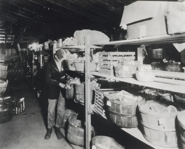 Man searching through cans of food piled in a wooden basket on a shelf in the distributing room of a community canning kitchen.
