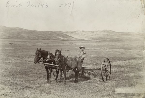 View across field towards a young man sitting on a horse-drawn dump rake pulled by a team of horses. In the background are hills.