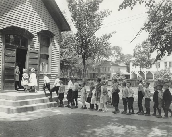 Group of young school children entering a school building single file, with arms folded behind their backs. The original caption reads: "Proper way of entering school room."