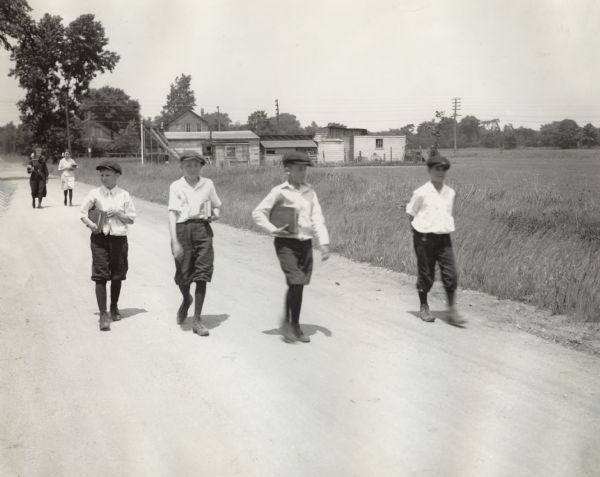 Four young boys walking on a dirt road in rural Cook County on their way to school. All are dressed similarly. Two older girls are also seen walking closely behind the group, carrying school books.