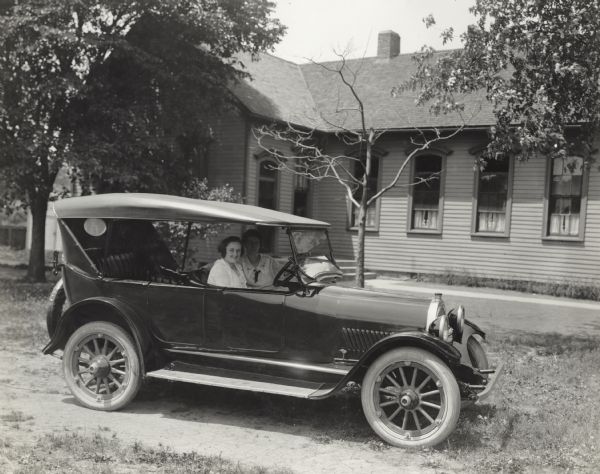 Two women seated in automobile outside school building(?). Original caption describes the women as Miss Sullivan and her assistant, Miss Brady, both rural teachers.