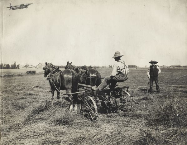 Man on a Reynolds hay tedder pulled by team of work horses. Another man stands supervising. In the far background beyond a fence are two separate farmsteads with barns, farmhouses and farm buildings.