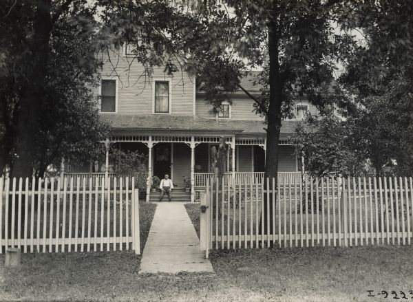 View from a street of a man seated on the front porch steps of a house. A picket fence and front walk are in the foreground.