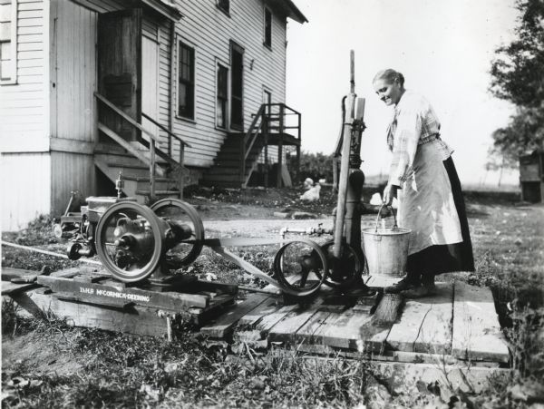 A woman using a 1.5 horsepower McCormick-Deering engine to pump water into a metal bucket outside a building at International Harvester's Hinsdale experimental farm (Harvester Farm).