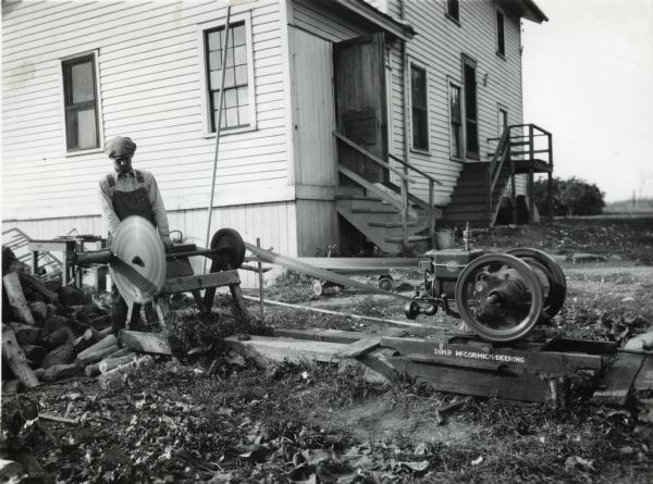 A man wearing overalls and a cap is using a saw powered by a 1.5 horsepower McCormick-Deering engine to cut wood on International Harvester's Hinsdale experimental farm (Harvester Farm). A building with sets of stairs leading to two entrances is behind him.