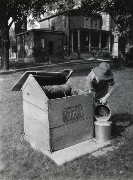 A woman wearing a dress and wide-brimmed hat is emptying water from one pail into another beside a hand-cranked well. The photograph was taken on the John Manier farm; the farmhouse is in the background.
