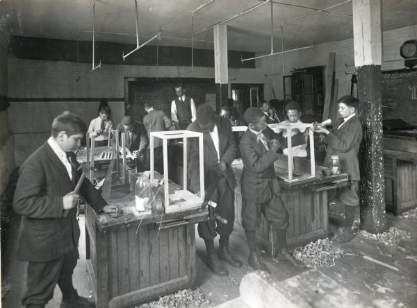 Boys use various tools to make fly traps in a classroom. A man, probably their instructor, stands in the background in front of a chalkboard.
