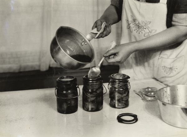 Woman preserving peaches in a kitchen at "Harvester Farm." She is pouring hot syrup from a pot over a spoon into glass jars filled with the fruit.