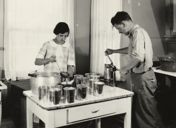 A woman and a man in a kitchen canning beans. The woman is scooping beans from a large pot with a ladle into cans, while the man uses a tabletop hand cranked sealer to put tops on the cans. The image was taken at "Harvester Farm."
