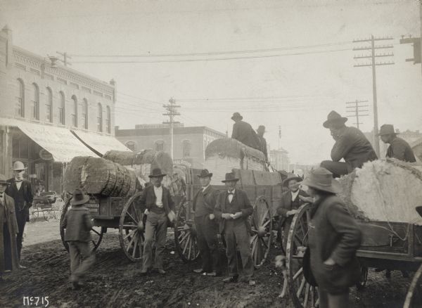 Group of men on a city street with bales of cotton loaded in horse-drawn wagons. One of the wagons is a Springfield Wagon.