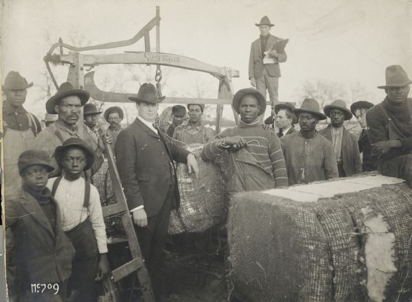 Group of men and young boys are gathered outdoors while weighing bales of cotton on a large hanging scale manufactured by Smith.