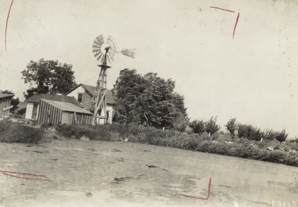 View over a pond of several farm buildings surrounded by trees and a windmill.