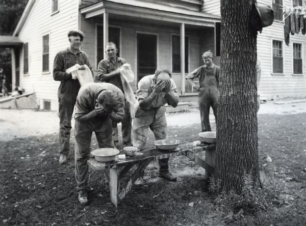 Men washing their faces with water in metal bowls set on a wooden bench outside a farmhouse. Two men are standing behind them, drying their hands on towels.