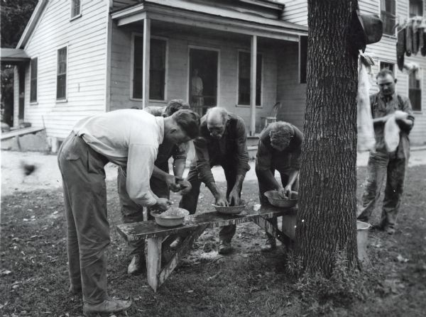 A group of men washing their hands in water bowls set on a wooden bench outside a farmhouse. A man is standing nearby while drying hands on a towel and a woman is looking on from behind a screen door of a farmhouse in the background.