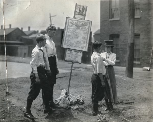 Three boys and a woman standing near several signs advertising a war garden. The signs read (from top to bottom): "Every Boy Can Feed a Soldier," "Registered War Garden Under Protection of State Council Defense. Feed Yourself. A War Garden Will Do It. Be a Soldier of the Soil. Exempt No Land," "To destroy the food supply is to give comfort to the enemy. Food Will Win the War," and "War Garden. Protected and Registered. No Loyal American Will Steal or Destroy This Crop."