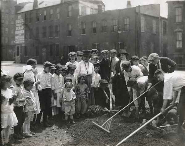 A group of children and women standing in what is probably a "war garden" in an urban area. Several commercial buildings are in the background, one painted with a sign reading: "H.L. Siegel. Scrap Rubber and Metal."