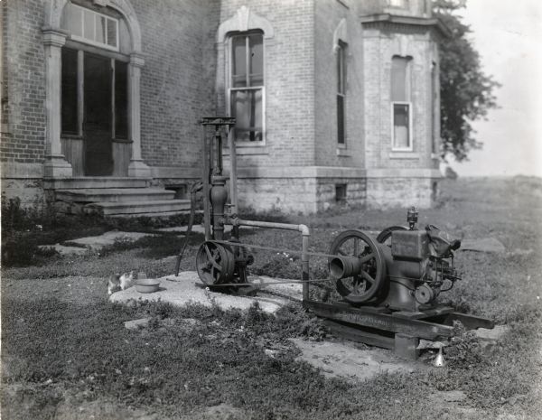 An International stationary engine powers a water pump outside a brick building, possibly a school. There is a cat on the left near the pump.