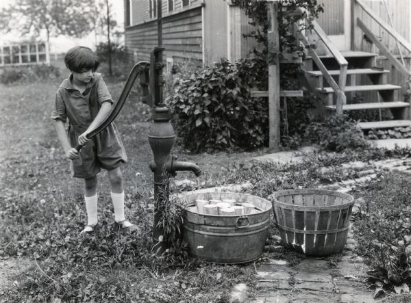 Young girl identified as Jewel Keller using a water pump outside a farm house to cool freshly canned corn.