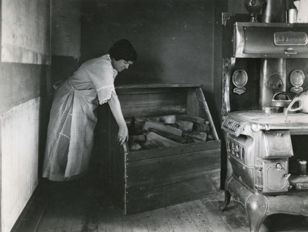 A woman moving a box full of firewood from the corner of the kitchen on the E.A. Bancroft farm. A woodstove is on the right.