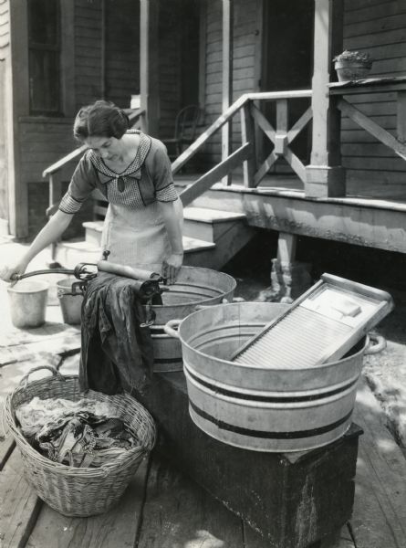 A girl is using a hand-crank clothing wringer and washboard to do laundry outside a farmhouse on the John Eshe farm.