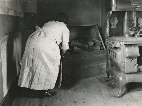 A woman moving a box of firewood from the corner of the kitchen on the farm of E.A. Bancroft. A woodburning stove is the right.